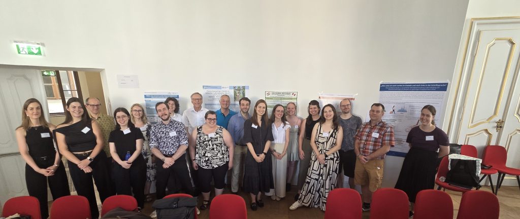 Gruppe von Personen, die in einem Raum stehen, umgeben von Plakaten. Sie tragen verschiedene Kleidung und posieren für ein Gruppenfoto.