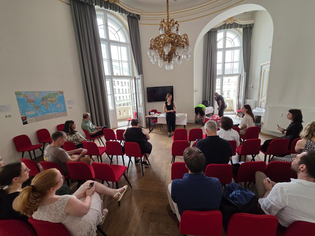 Ein Raum mit roten Stühlen, in dem eine Präsentation stattfindet. Im Vordergrund steht eine Person, die spricht, während die Zuhörer aufmerksam zuschauen. Im Hintergrund sieht man einen Bildschirm und eine offene Tür mit Fenstern.
